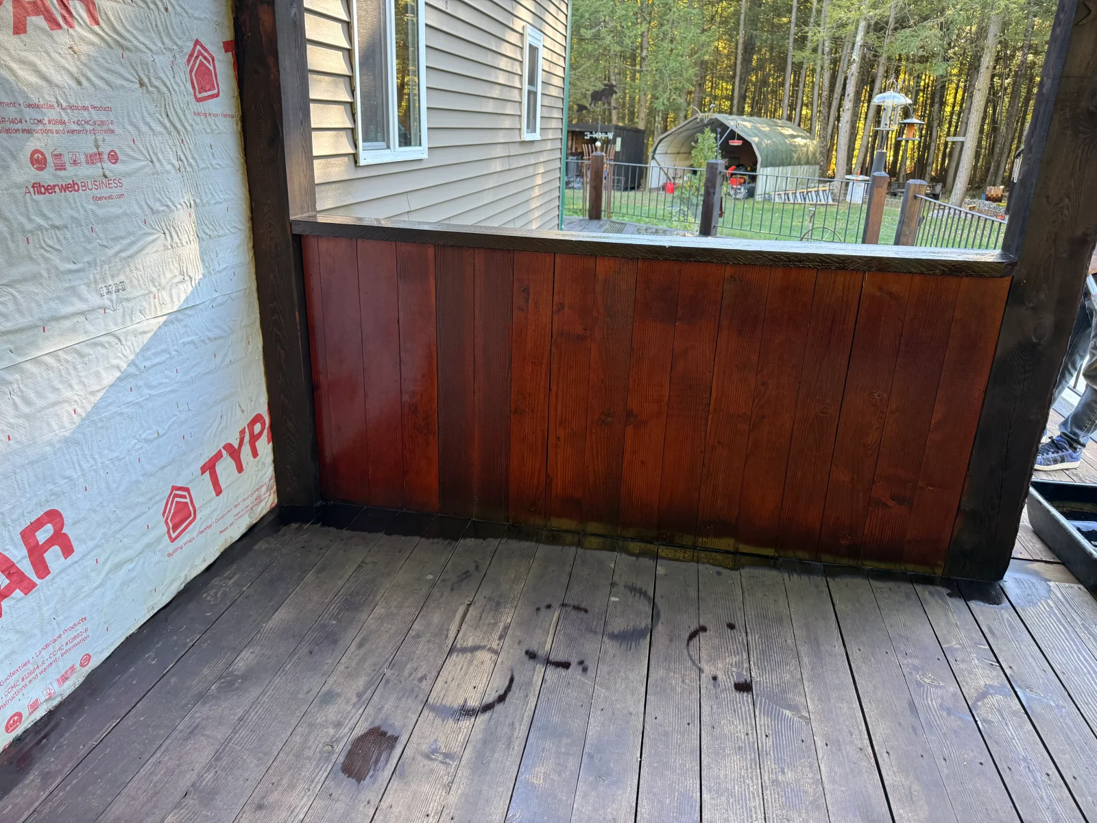 Vertical wood board privacy wall with reddish-brown stain on covered porch, dark stained top rail and posts, weathered deck boards.