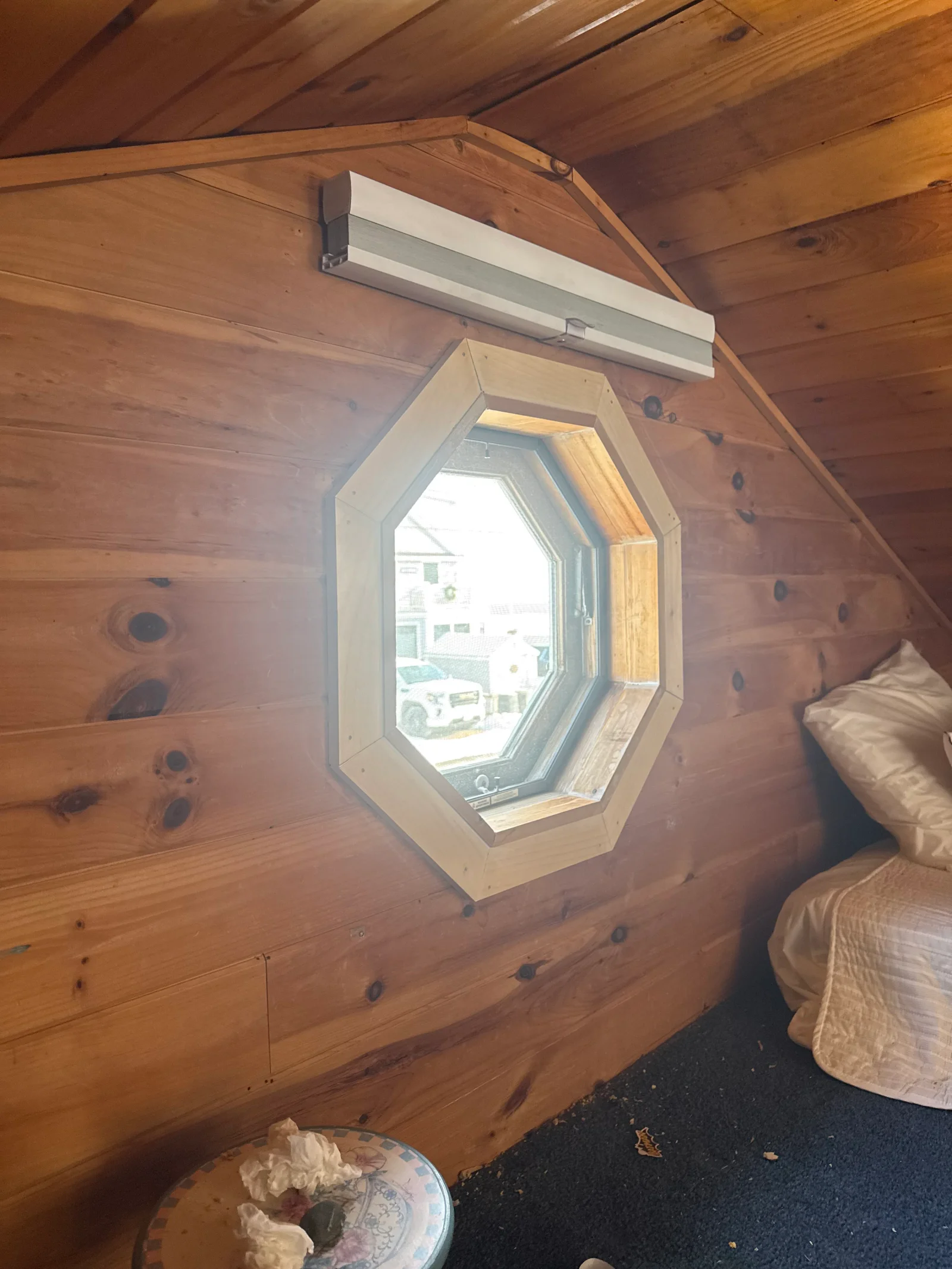 Octagonal window with light wood trim set in knotty pine paneled wall under sloped ceiling