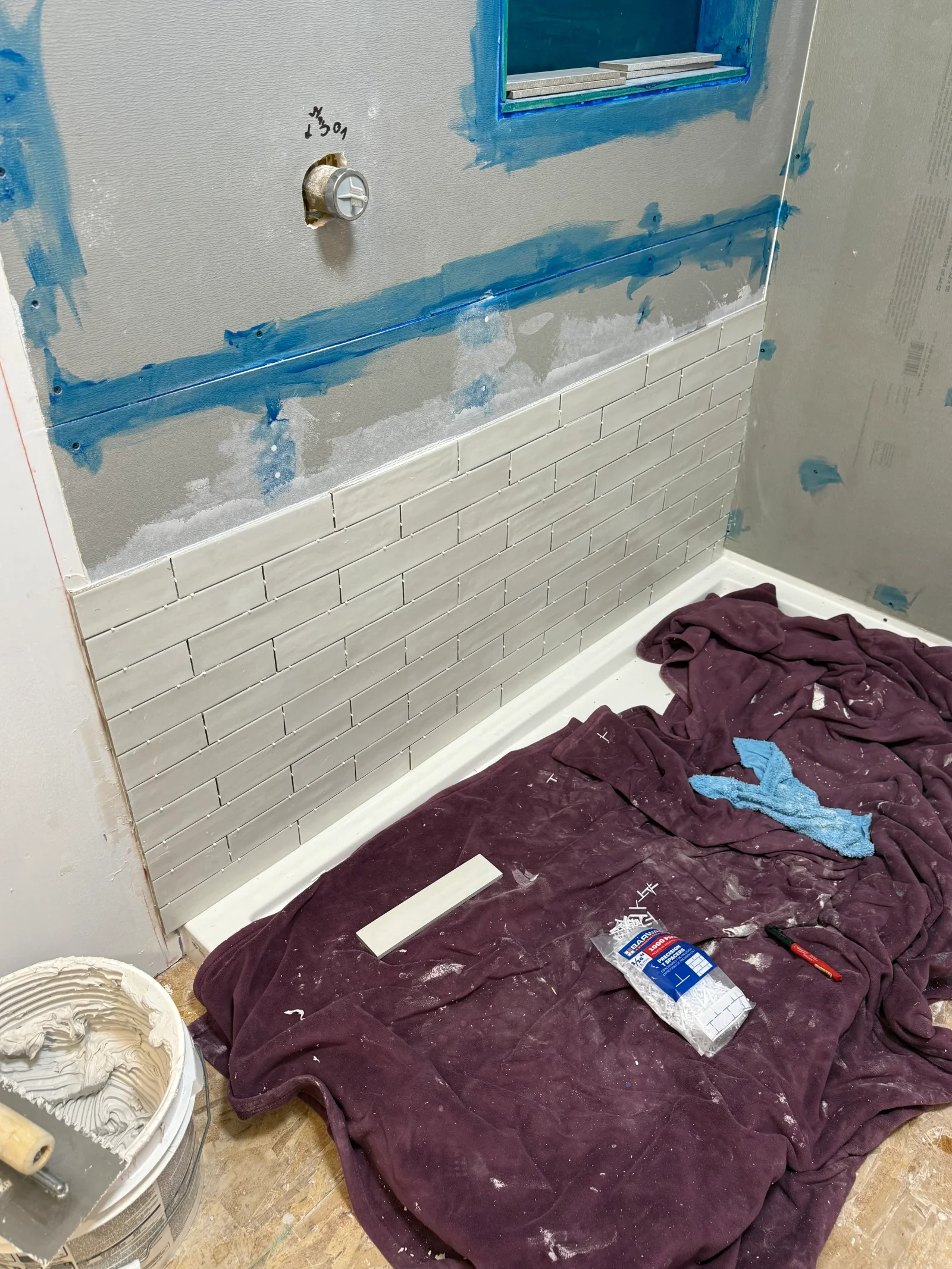Light gray rectangular wall tiles in running bond pattern being set above a white shower pan with blue tape and thinset mortar bucket