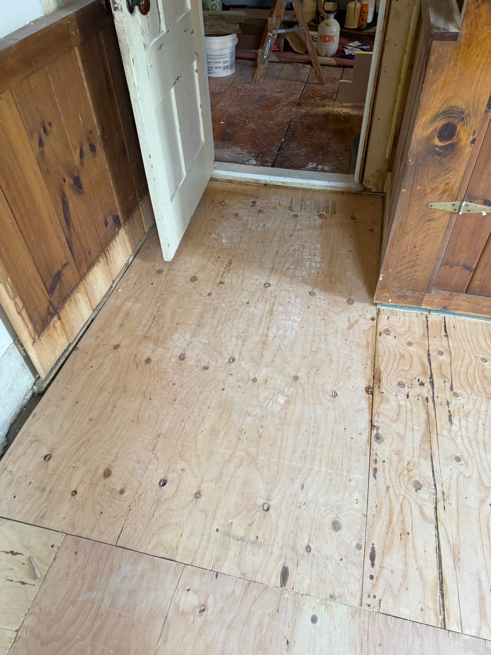 Plywood subfloor panels being installed over joists beside knotty pine wainscoting and an original painted door