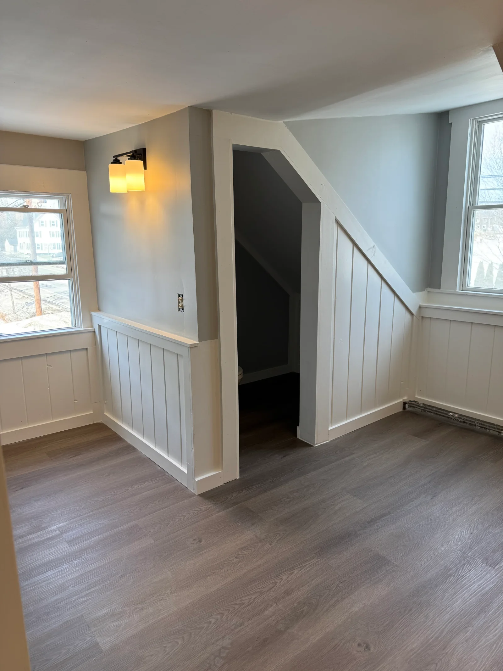Interior room with white vertical board-and-batten wainscoting, gray plank flooring, and frosted glass wall sconce beside a cased stair opening