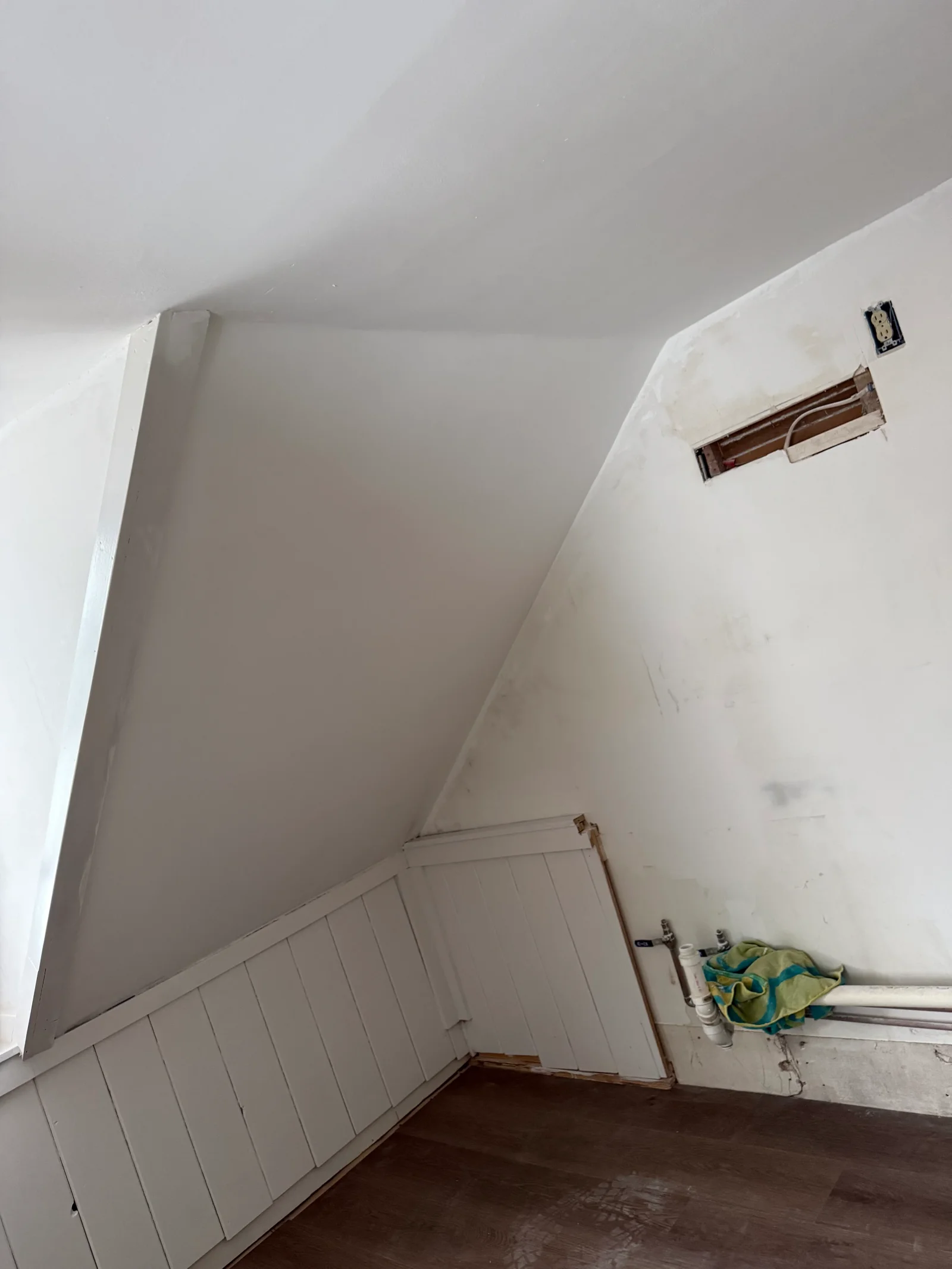 Attic room with white painted board-and-batten knee wall, sloped ceiling, and exposed outlet and plumbing during renovation