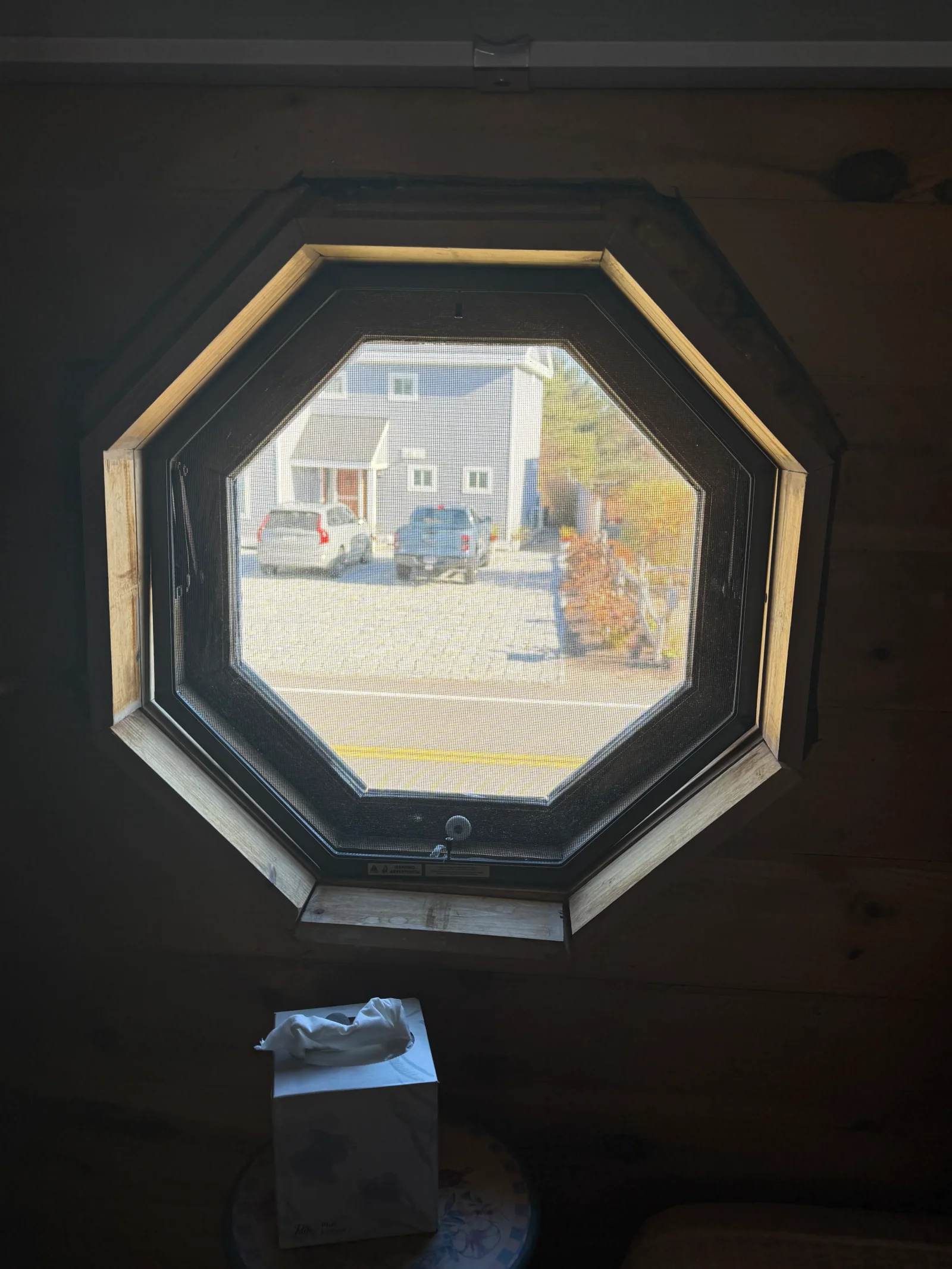 Octagonal window with dark frame and screen, surrounded by light natural wood trim in a wood-paneled wall