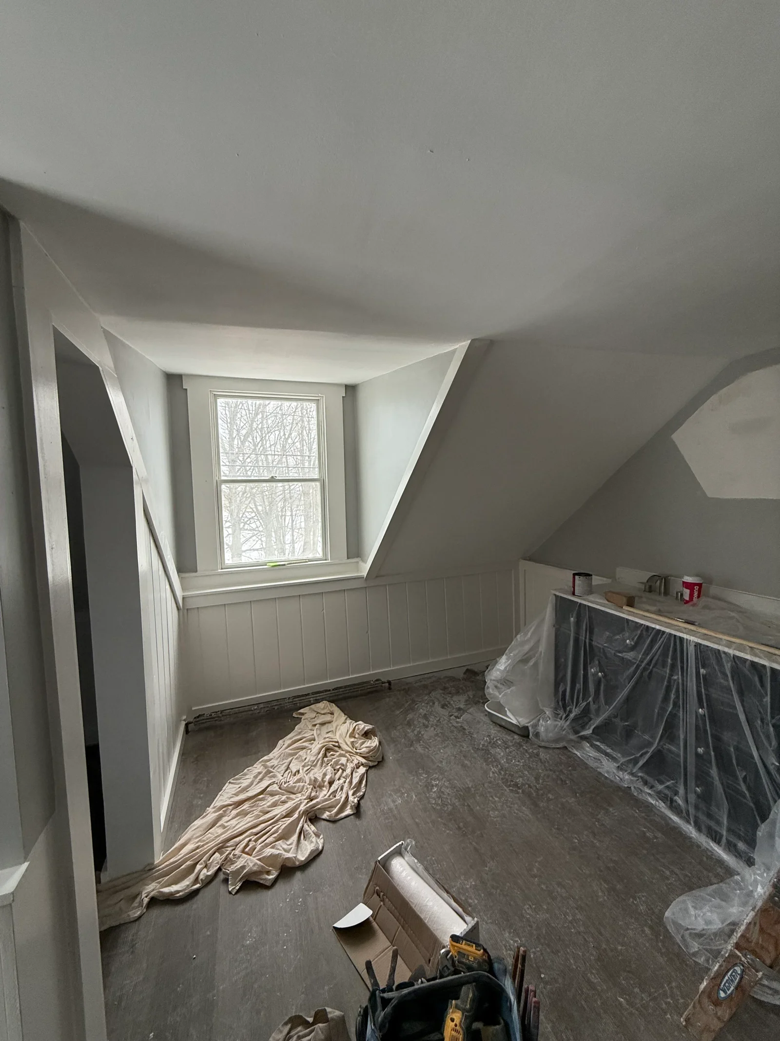 Attic room with gray painted sloped walls, white beadboard wainscot, and plastic-wrapped furniture during painting work