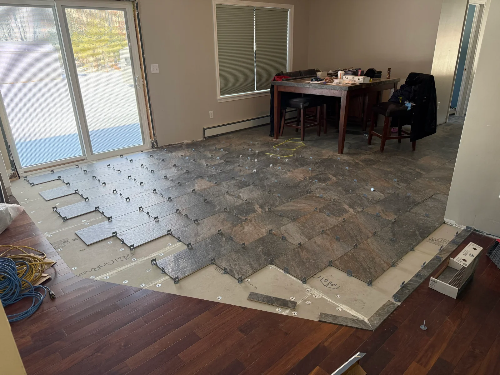 Slate-look porcelain floor tiles being laid over uncoupling membrane with leveling clips, bordering dark hardwood flooring