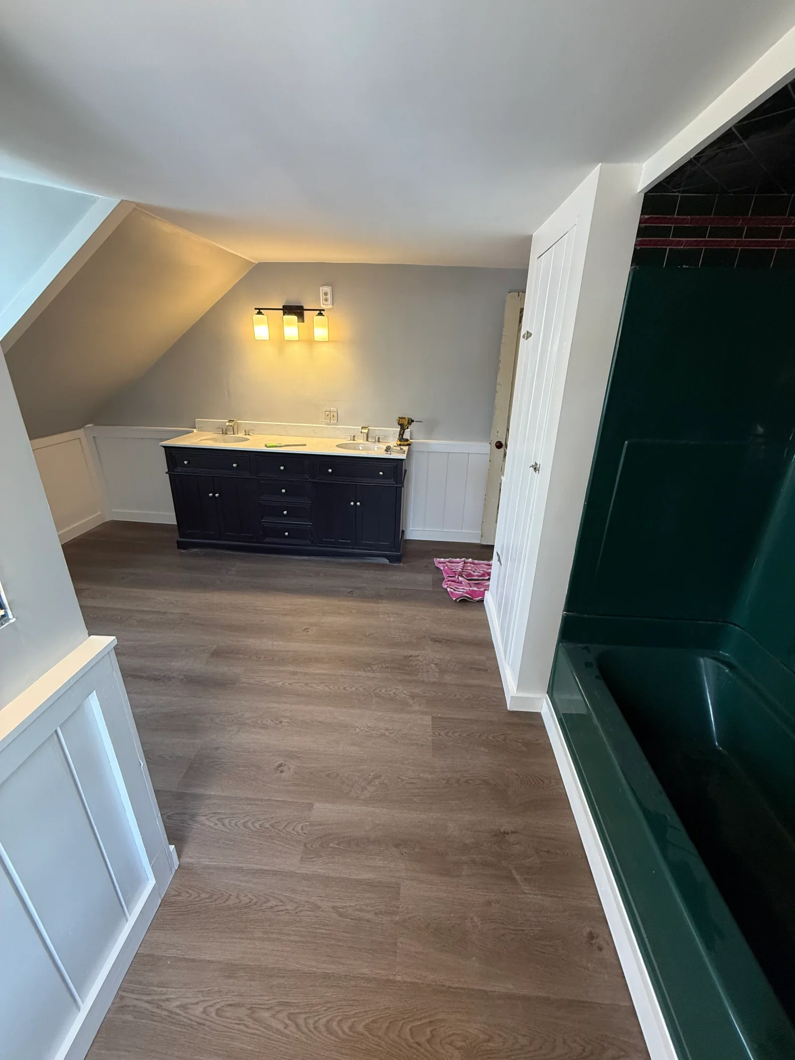 Attic bathroom with navy double vanity, white beadboard wainscoting, wide-plank vinyl flooring, and green tub surround