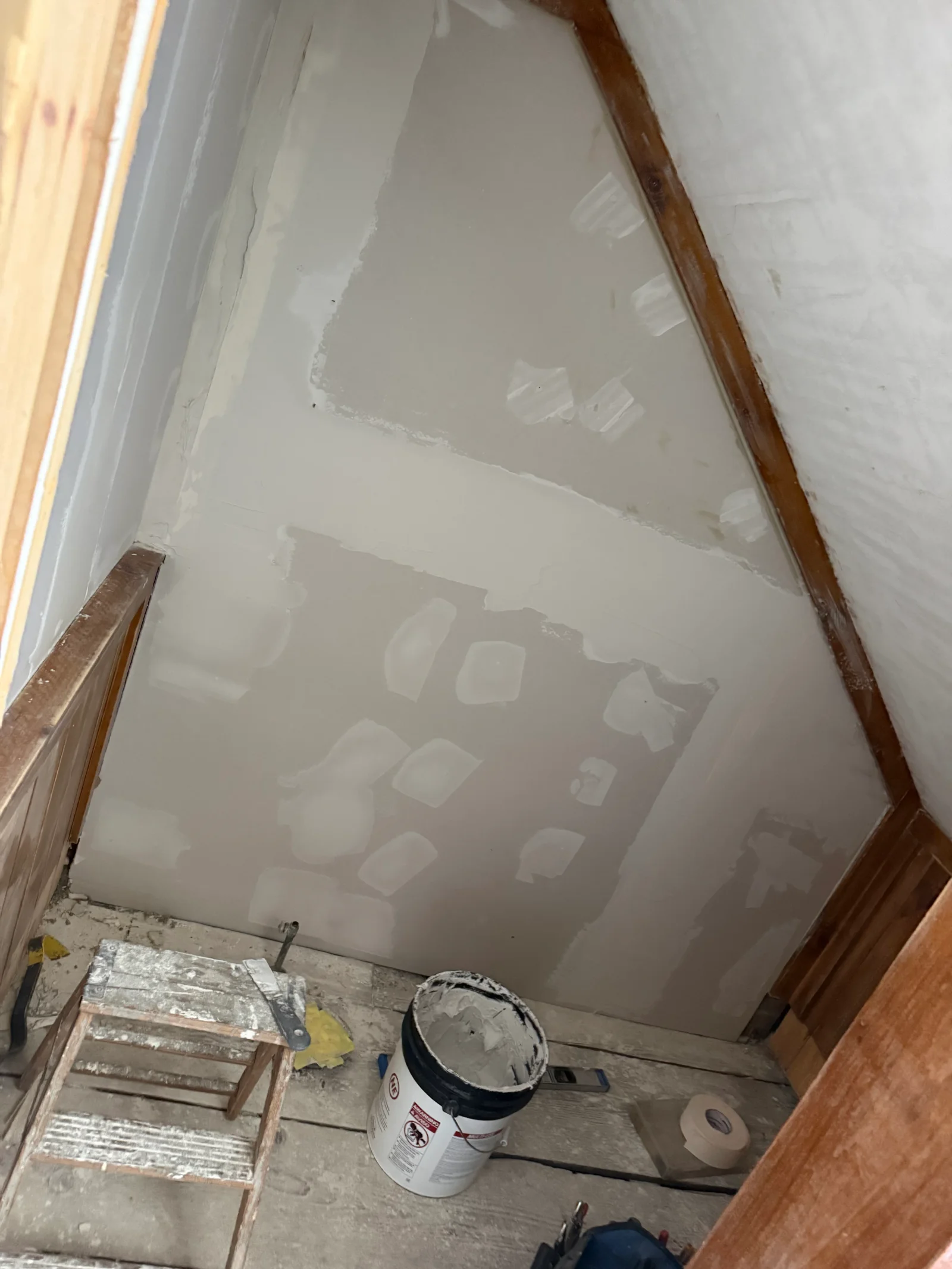 Sloped ceiling with drywall patched and skim coated with joint compound, wood trim and bucket of compound on floor
