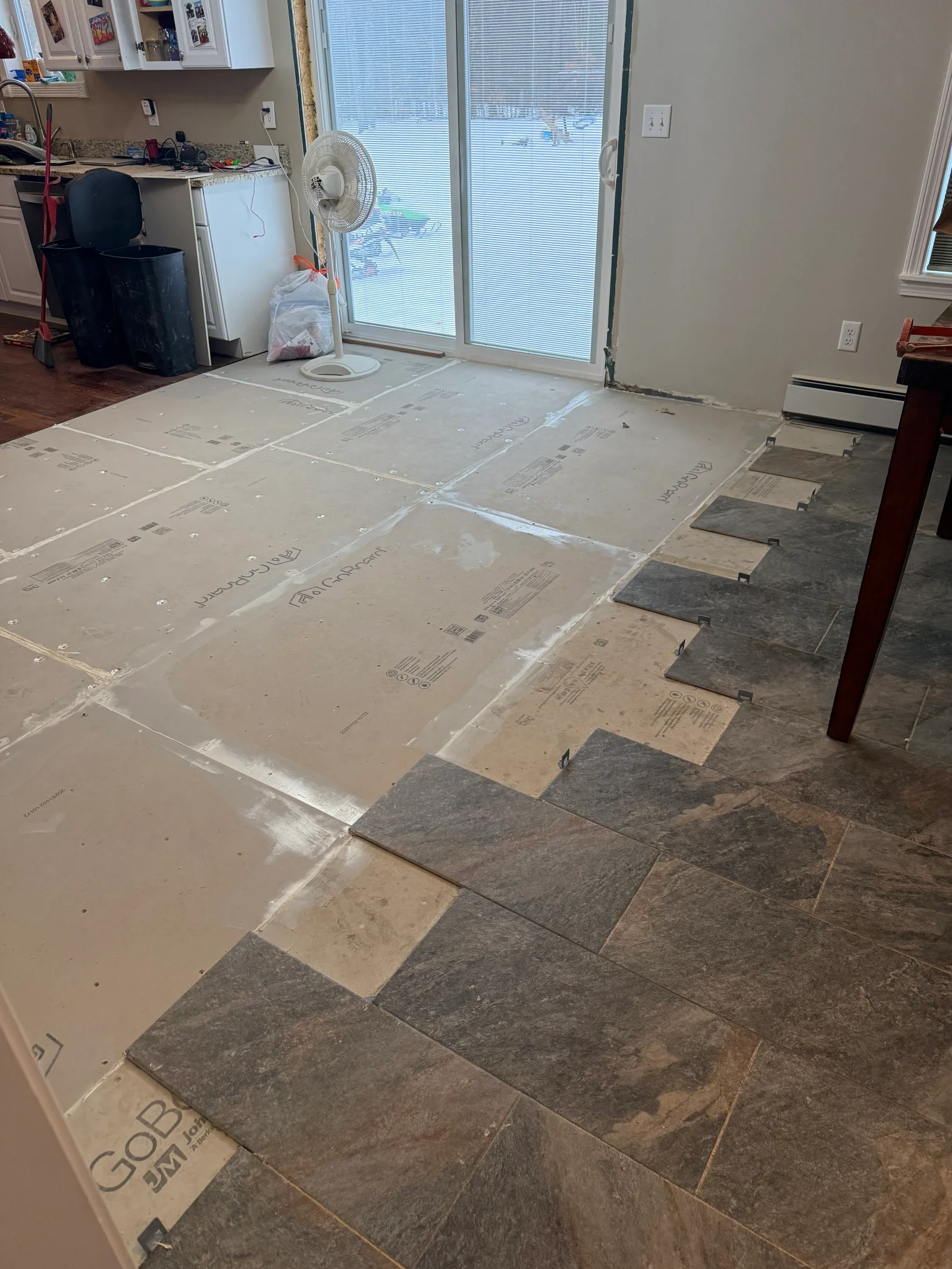 Cement board underlayment with dark slate-look floor tile partially installed in a kitchen, with leveling clips