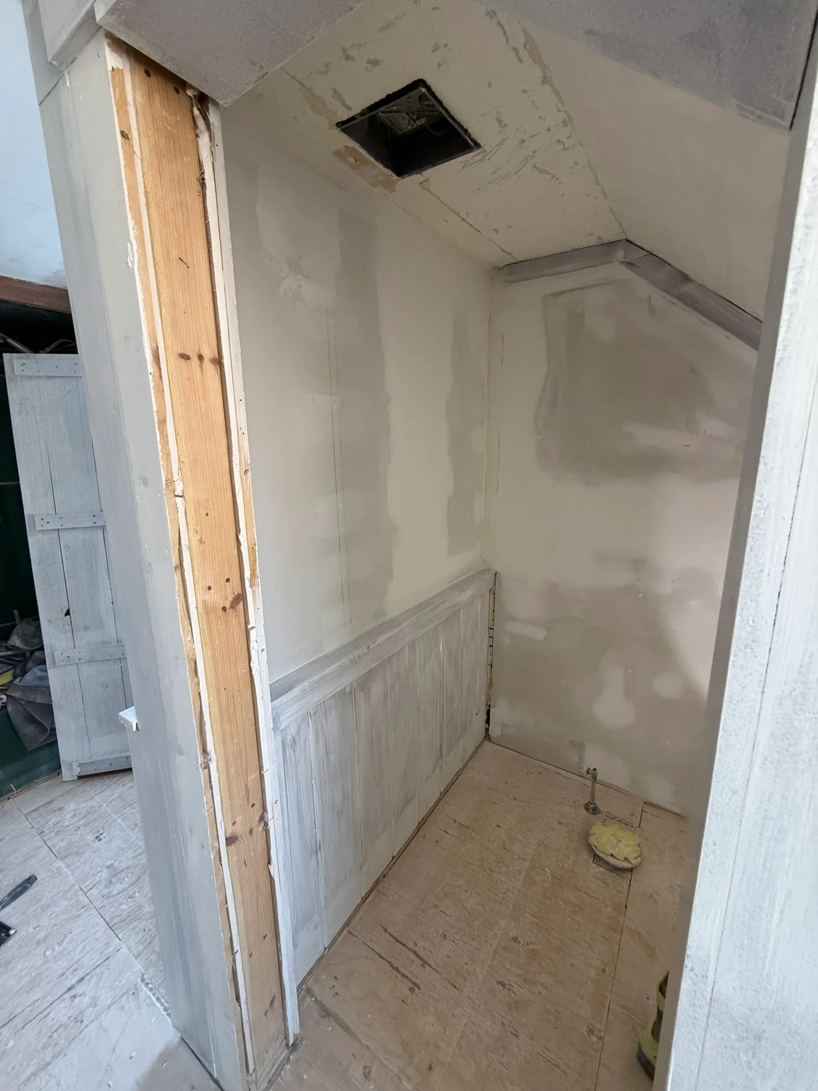 Alcove under renovation with sloped ceiling, joint compound walls, distressed painted wainscot, and plywood subfloor