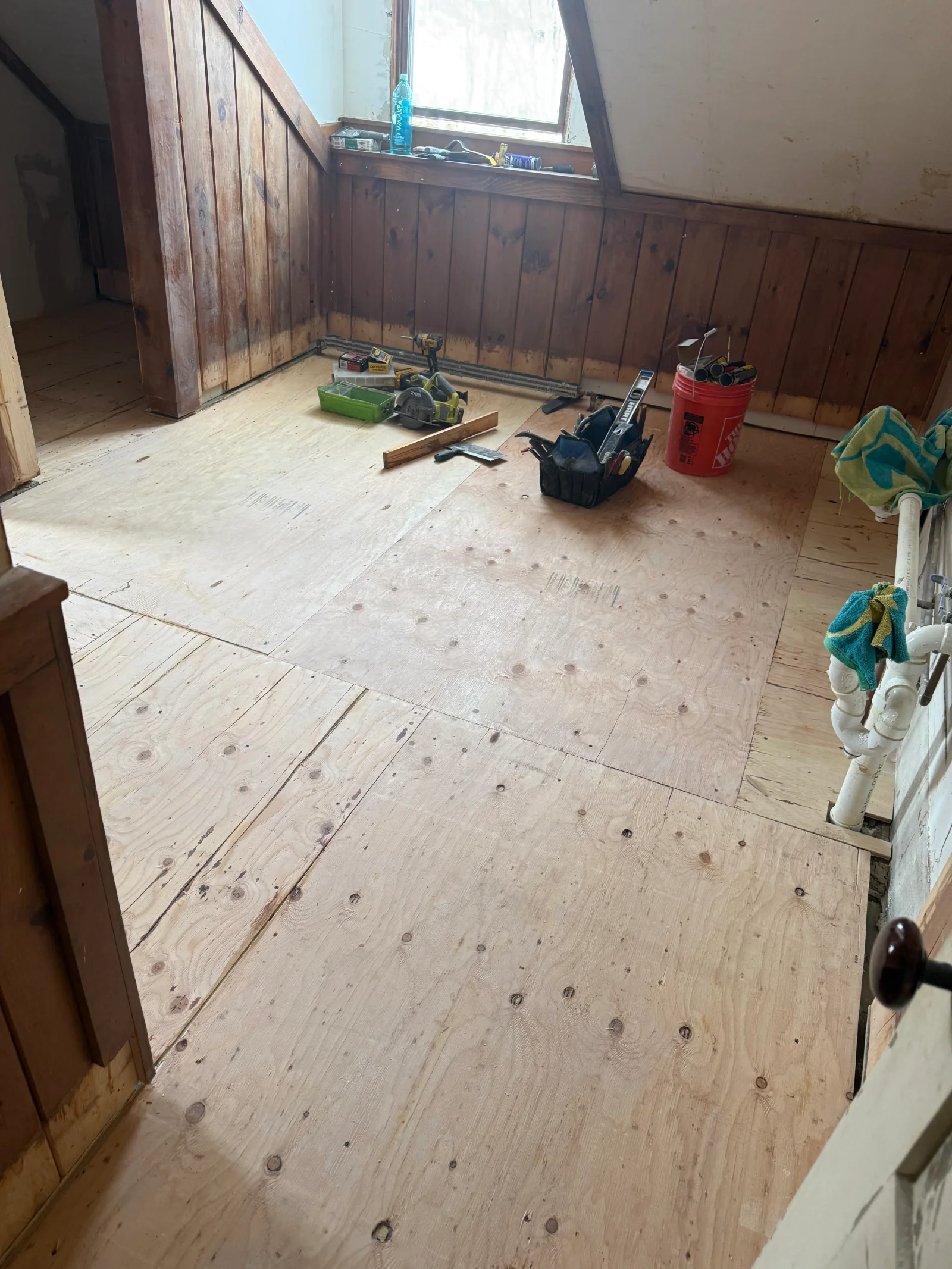 Attic room with plywood subfloor sheets being installed over old boards, exposed wood paneling, and carpentry tools on the floor