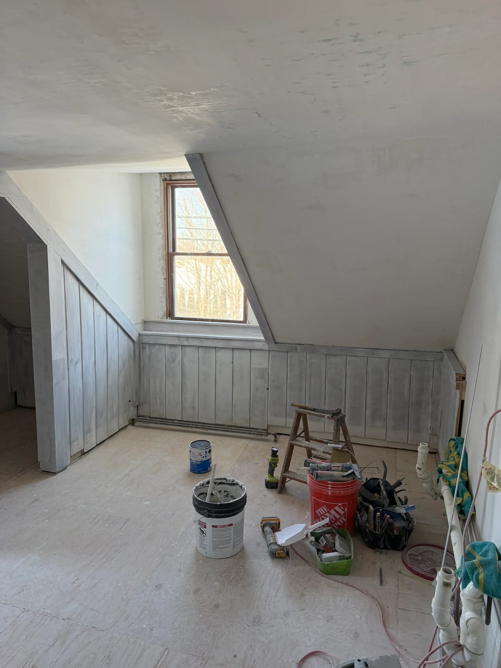 Attic room with gray vertical board paneling on knee walls, sloped ceiling, and exposed plywood subfloor during renovation