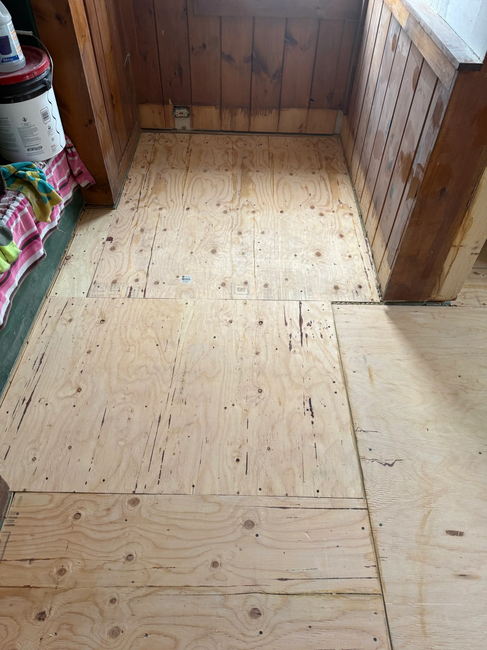 Plywood subfloor panels being installed over an open floor with wood-paneled walls and fastened seams