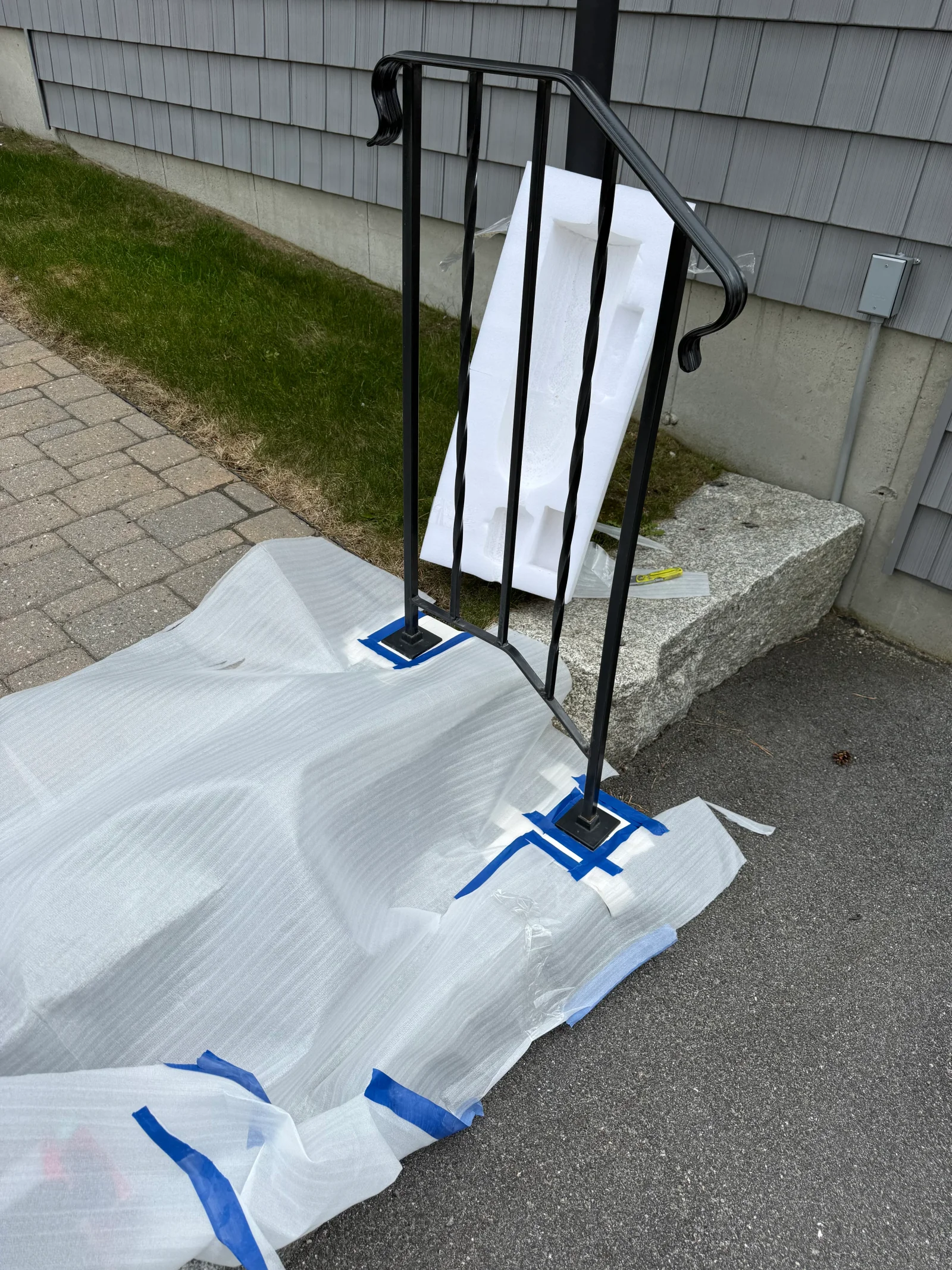 Black wrought iron handrail with twisted balusters and scrolled ends, base plates masked with blue tape and covered in white protective wrap