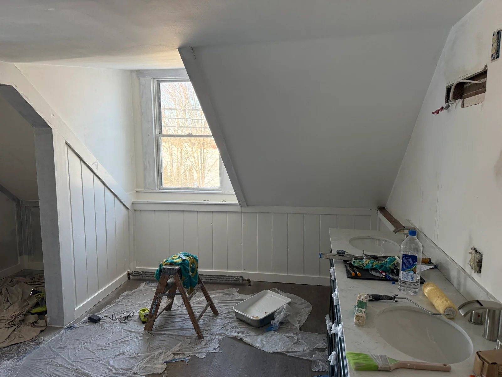 Attic bathroom with white painted vertical wainscoting, sloped ceiling, double vanity with marble-look countertop, and painting supplies on drop cloths