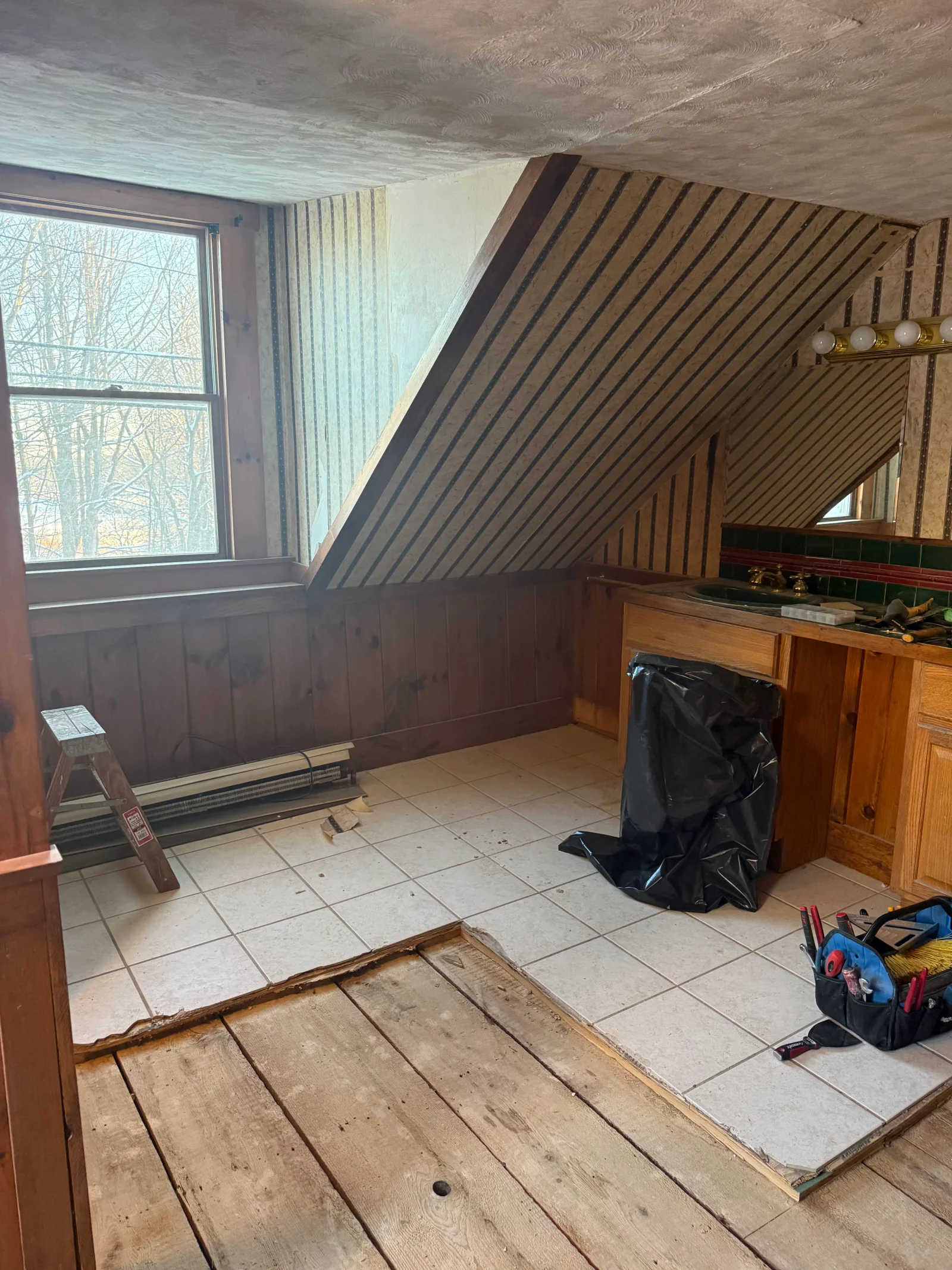 Partially demolished attic bathroom with pulled-up ceramic tile exposing wide wood plank subfloor, knotty pine wainscoting, and sloped striped wallpaper
