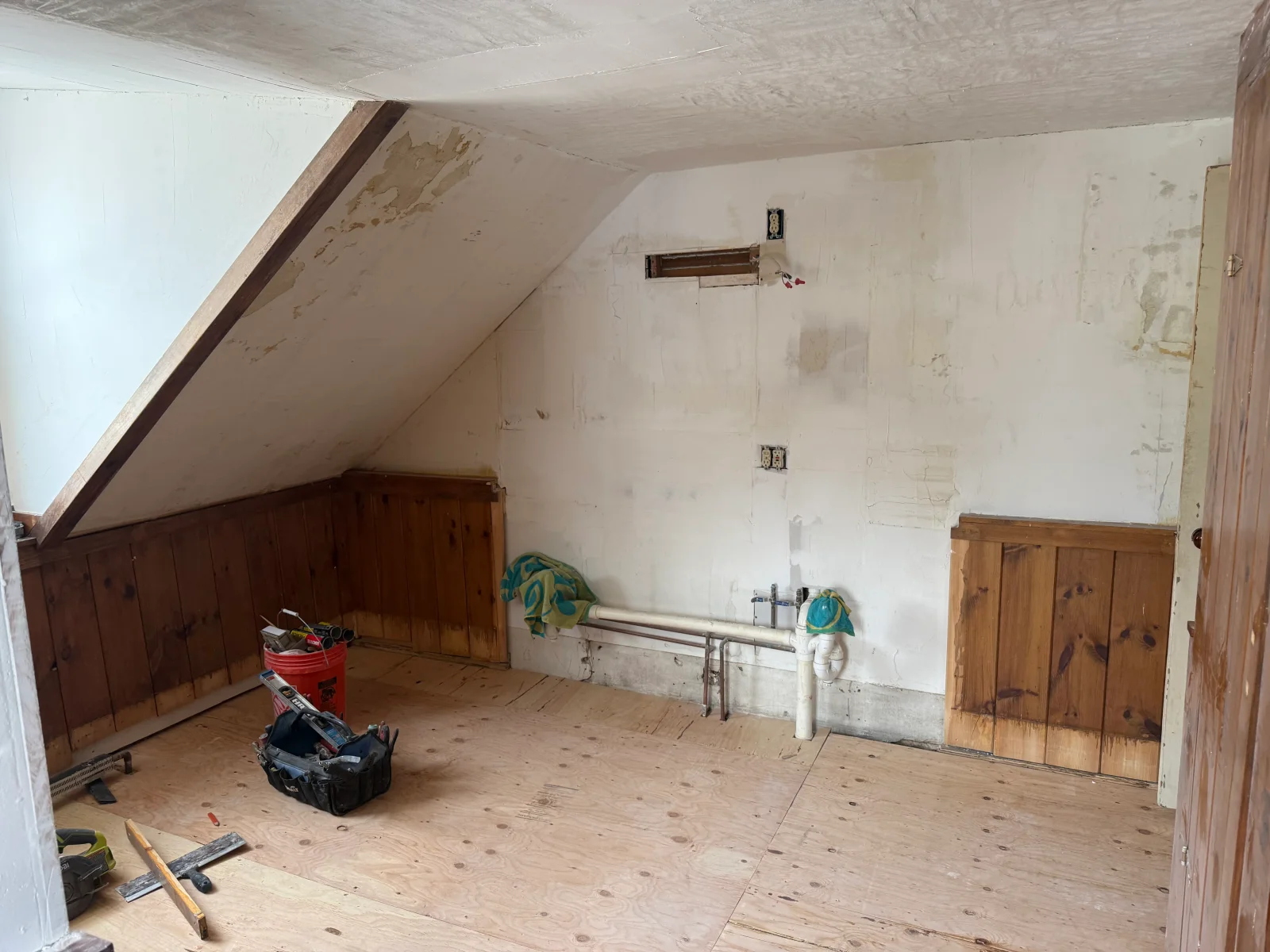 Attic room with sloped ceiling, plywood subfloor, wood wainscoting, exposed PVC drain and copper pipes