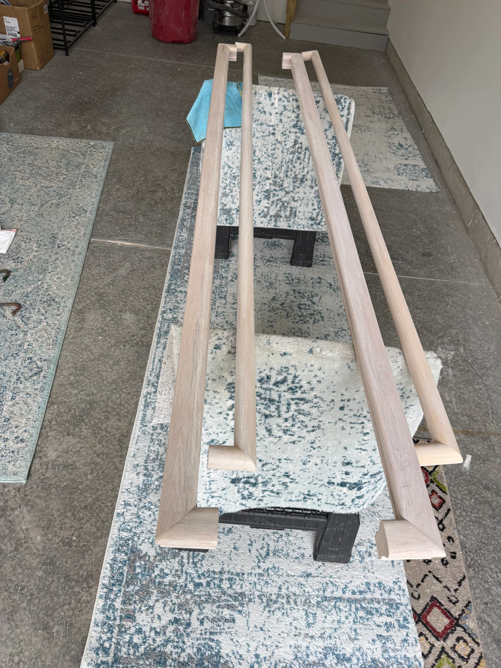 Pair of long light-colored wood bar door pull handles with angled mitered ends, laid out on a rug in a garage workspace